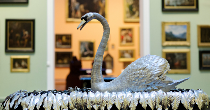Photograph of a detailed silver swan sculpture displayed in an art gallery with framed paintings on walls in the background. Sculpture features intricately crafted feathers and a long, curved neck, serving as a central decorative piece amidst classical artworks.